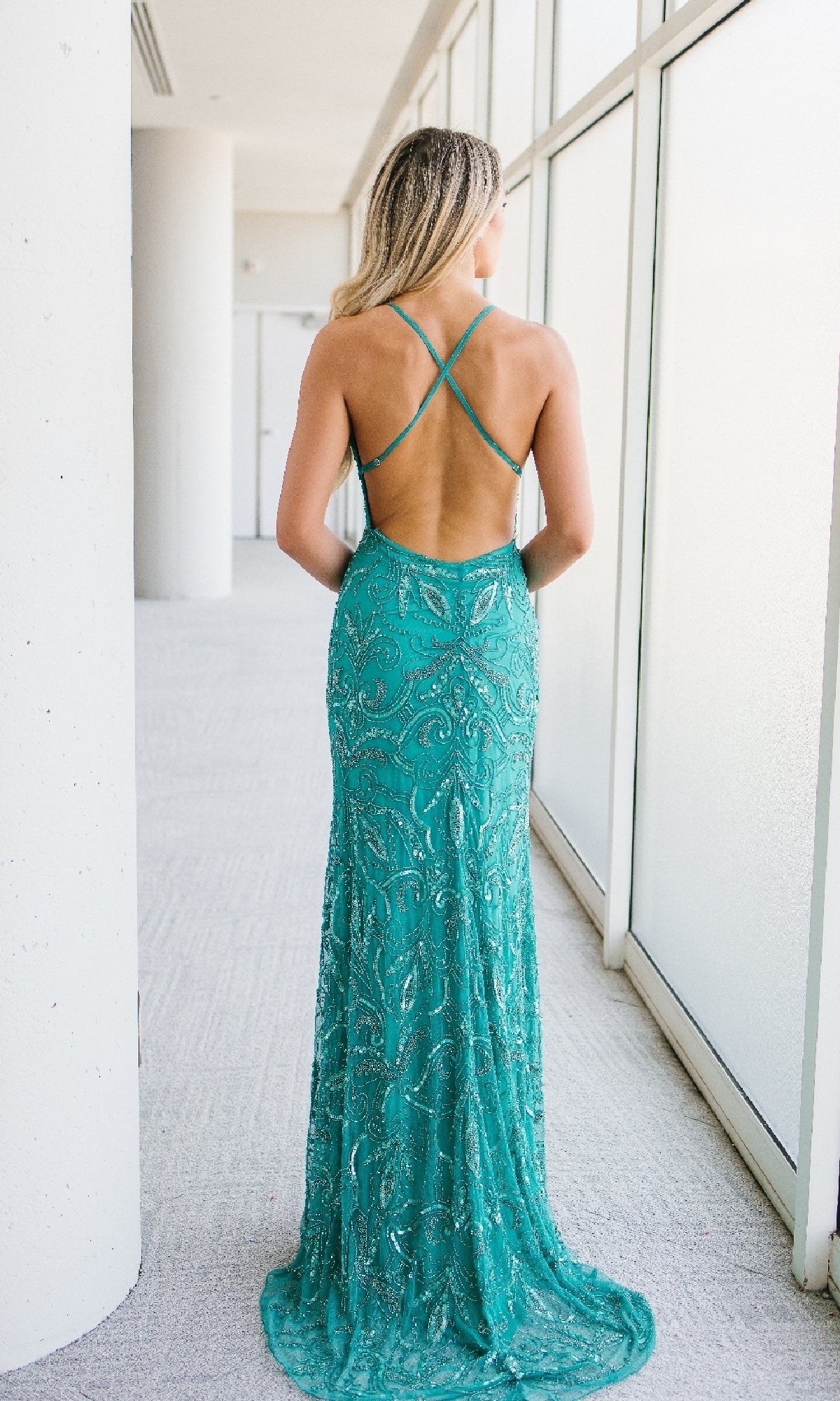 A woman stands indoors facing away, wearing the Long Sequin-Print Prom Dress with a strappy open back in teal—a stunning formal evening gown ideal for special occasions.